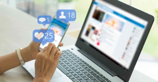 A hand holding a phone perusing social media. The computer is also open to a social media page